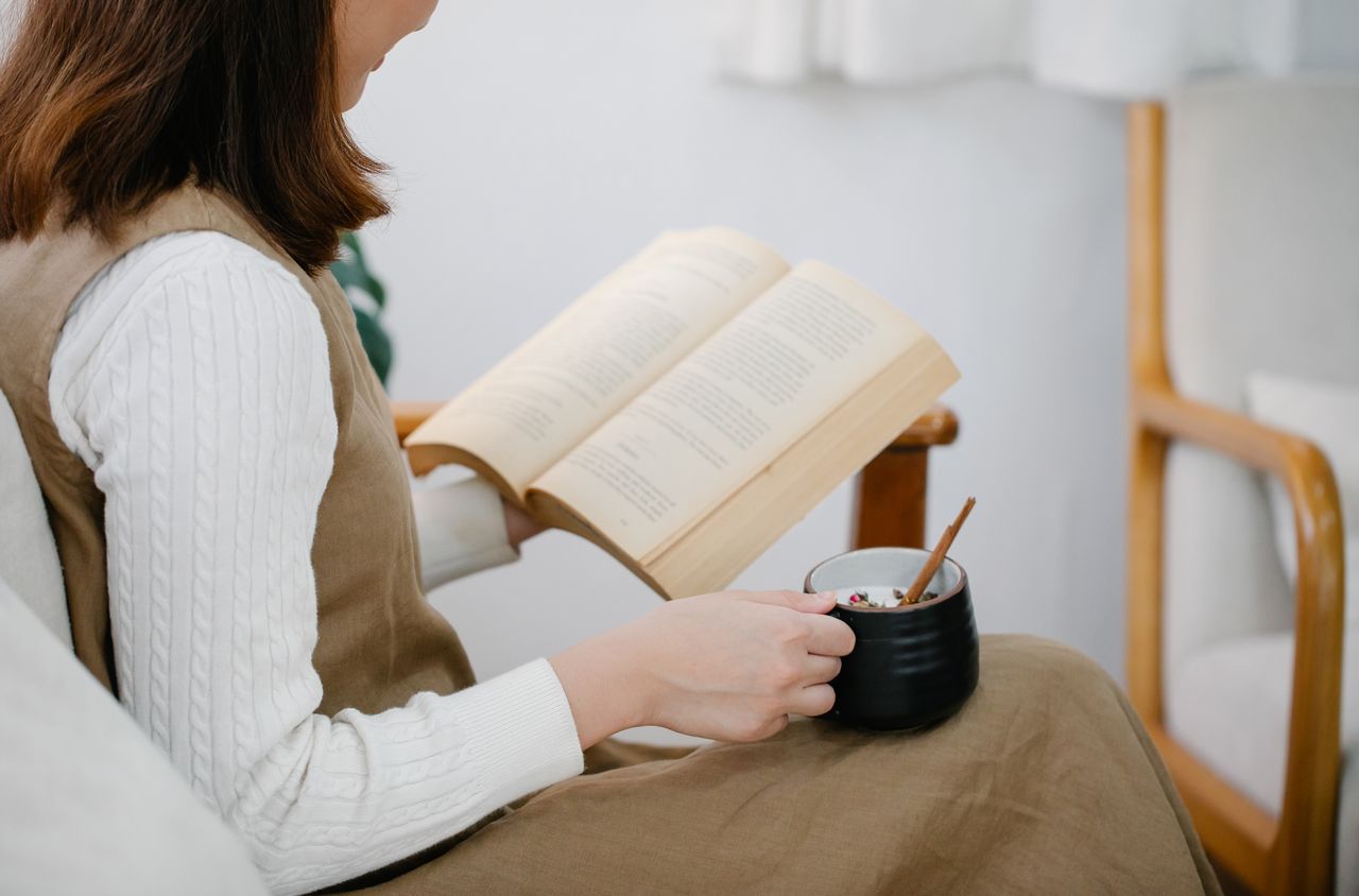 Woman sitting in an armchair reading a book and holding a cup of tea, in a peaceful indoor setting.